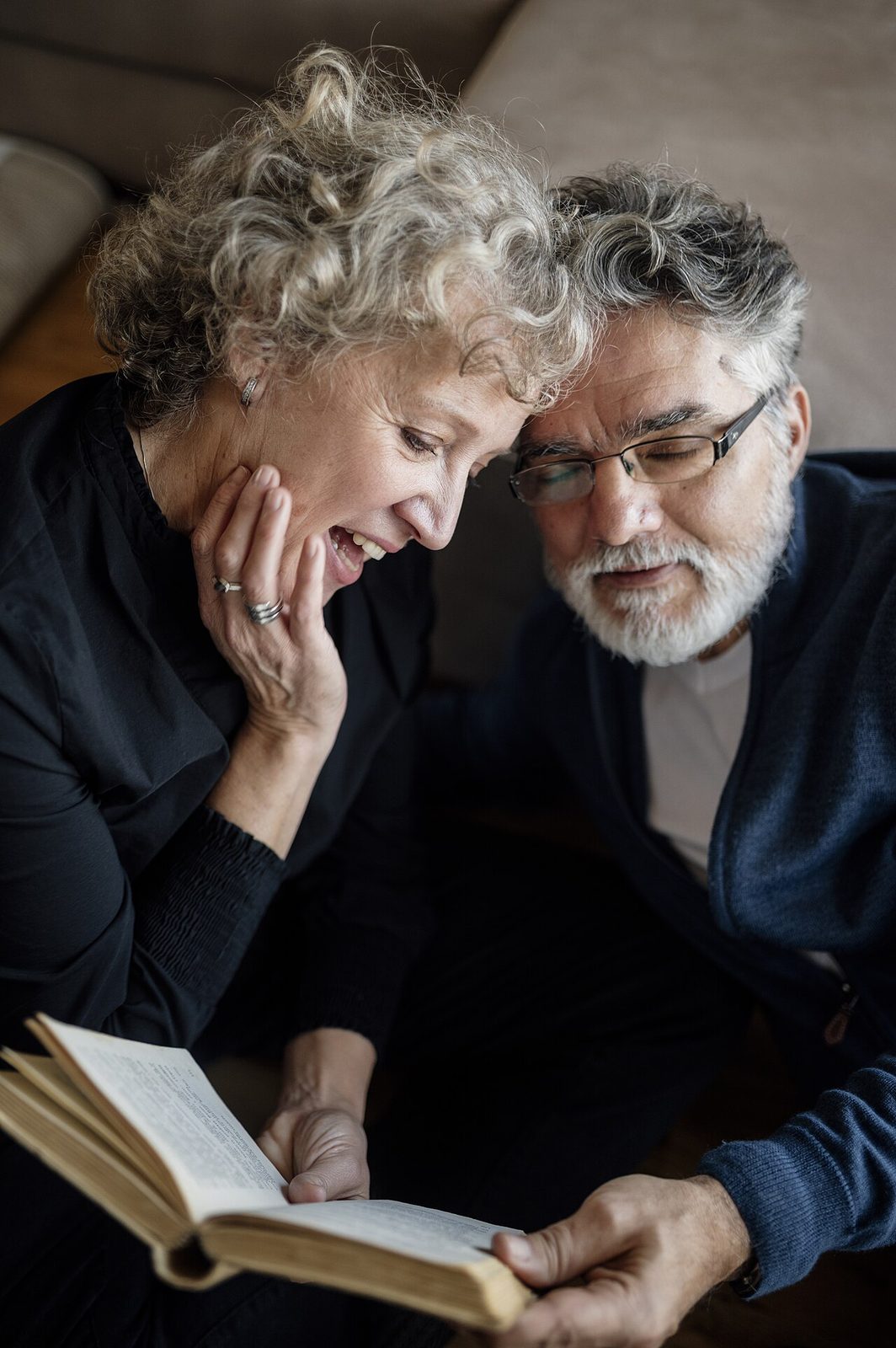 An elderly couple sitting together at home, one reading aloud while the other listens, a quiet afternoon light through the window.