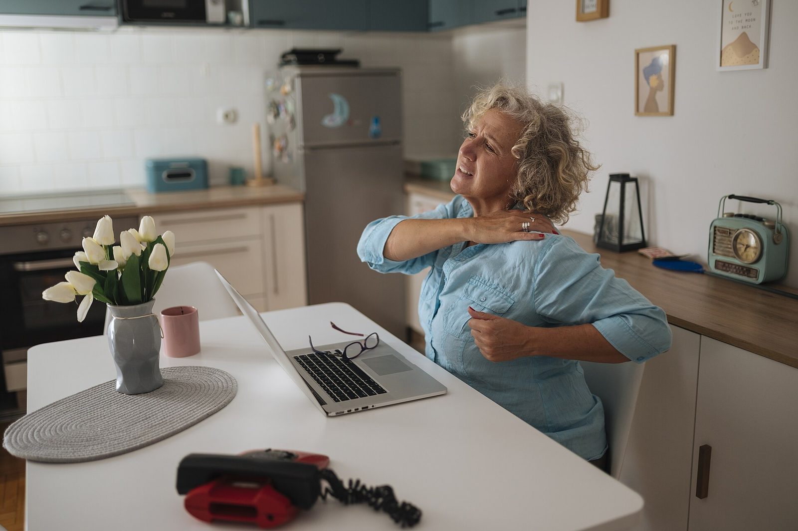 A senior woman working on a laptop at a wooden desk at home, a notebook open beside her, morning light.